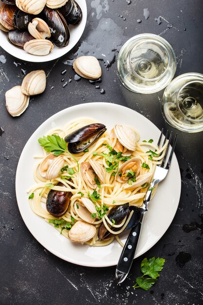 Vongole de espaguetis, pasta de marisco italiano con almejas y mejillones, en plato con hierbas y dos copas de vino blanco sobre fondo de piedra rústica.