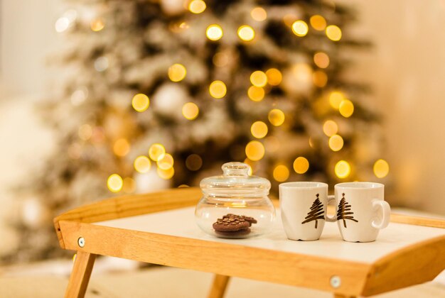 Vor dem Hintergrund der Weihnachtsbeleuchtung steht ein Holztablett auf dem Bett. Auf dem Tablett steht ein Glas mit Keksen und zwei Tassen