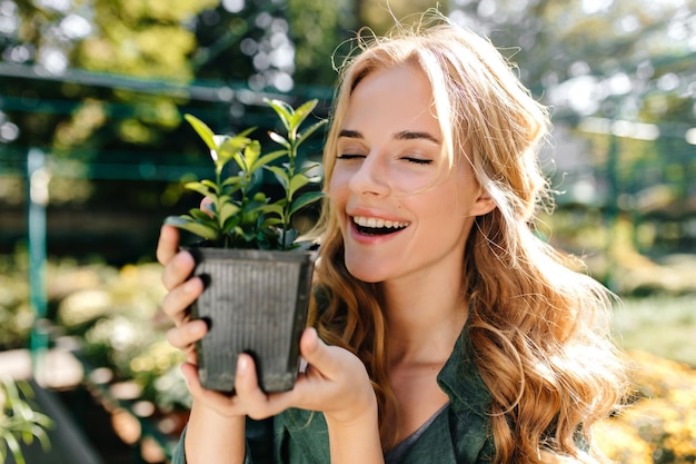 Vor dem Hintergrund einer großen Anzahl von Grüns und Pflanzen und Sonnenschein süßes Mädchen mit sanftem Make-up, das vor der Kamera posiert und einen Topf mit grüner Blume hält