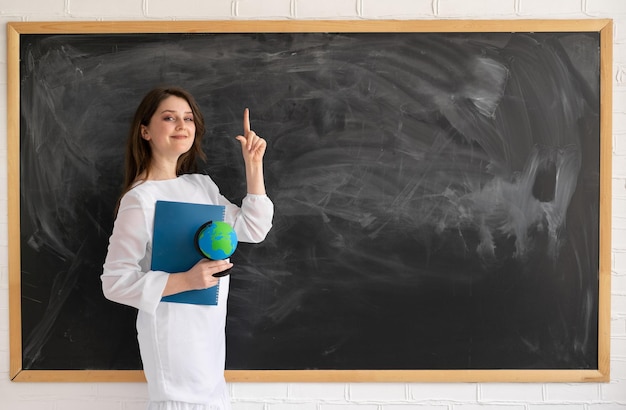 Vor dem Hintergrund einer sauberen Kreidetafel steht eine kaukasische Dame und hält ein Notizbuch in den Händen, der Zeigefinger zeigt nach oben Zurück zum Schulkonzept Lehrer oder Tutor