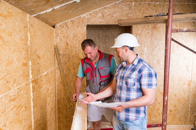 Vorarbeiter mit weißem Schutzhelm mit Bauplänen, der Arbeiter im unfertigen Rahmen des neuen Hauses überwacht