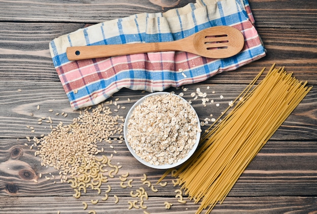 Vorbereitung des morgendlichen Frühstücks. Eine Schüssel mit rohem Haferflocken, Nudeln, Holzlöffel und einem Handtuch auf einem Holztisch. Der Kochvorgang. Draufsicht.