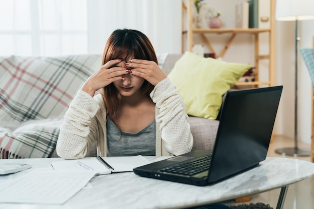 Vorderansicht Asiatisches Mädchen, das sitzt und ihren Kopf in den Händen vergräbt, ist besorgt über Schulden, während sie mit einem Laptop im Wohnzimmer zu Hause Steuern einreicht.