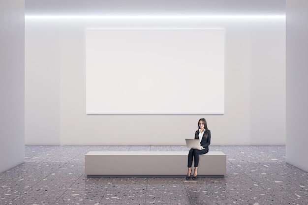 Vorderansicht auf leeres weißes Plakat mit Platz für Ihr Logo auf hellem Wandhintergrund in geräumigem Galeriesaal mit Geschäftsfrau, die Laptop benutzt und auf Steinbank auf Betonboden sitzt