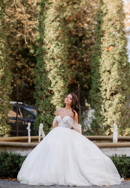 Vorderansicht der eleganten brünetten Braut steht auf dem Hintergrund des Parks mit einem Brunnen