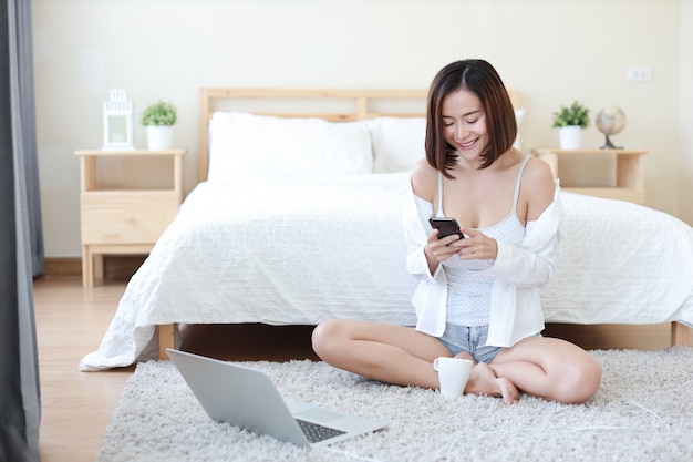 Vorderansicht der erwachsenen freiberuflich tätigen asiatischen Frau im weißen Hemd, das an Computer und Handy im Schlafzimmer mit glücklichem lächelndem Gesicht arbeitet