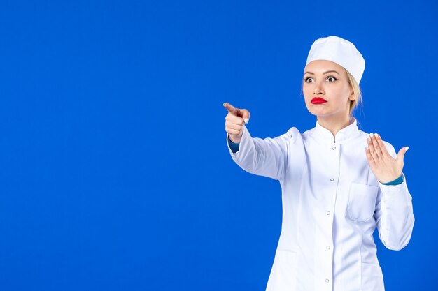 Vorderansicht der jungen Krankenschwester im medizinischen Anzug, die auf etwas an der blauen Wand zeigt