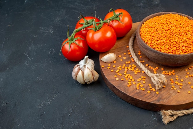 Vorderansicht der roten Linsen in einer braunen Schüssel Seilknoblauch auf hölzernen runden Bretttomaten mit Stielen auf der rechten Seite auf schwarzem Hintergrund