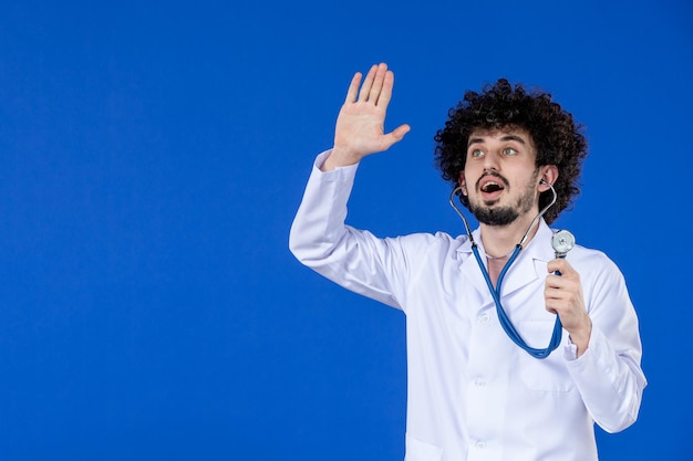 Vorderansicht des jungen männlichen Arztes im medizinischen Anzug mit Stethoskop auf blauer Oberfläche