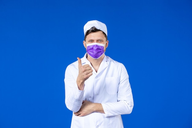 Vorderansicht des lächelnden männlichen Arztes im medizinischen Anzug und in der lila Maske auf Blau