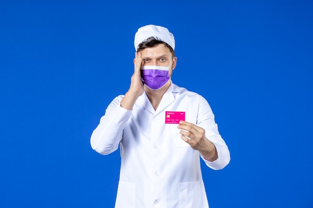 Vorderansicht des männlichen Arztes im medizinischen Anzug und in der lila Maske, die Kreditkarte auf Blau hält