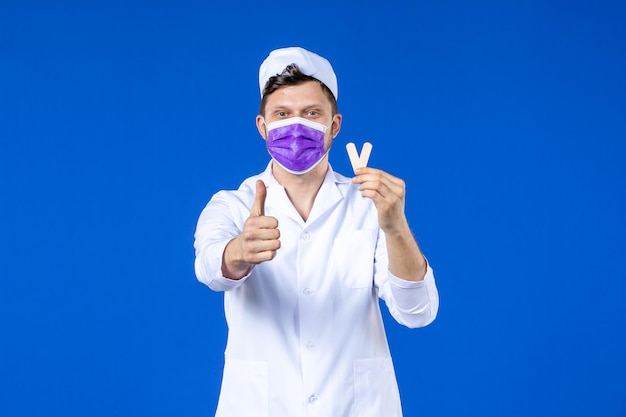 Vorderansicht des männlichen Arztes im medizinischen Anzug und in der Maske, die kleine medizinische Flecken auf Blau halten