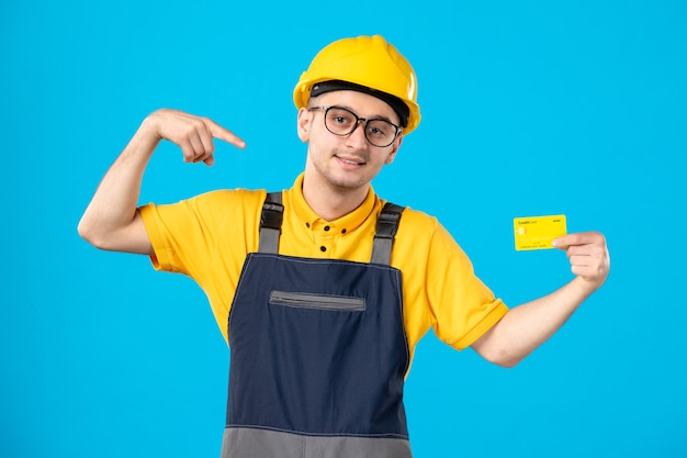 Vorderansicht des männlichen Baumeisters in der Uniform und im Helm mit Kreditkarte auf blauer Wand