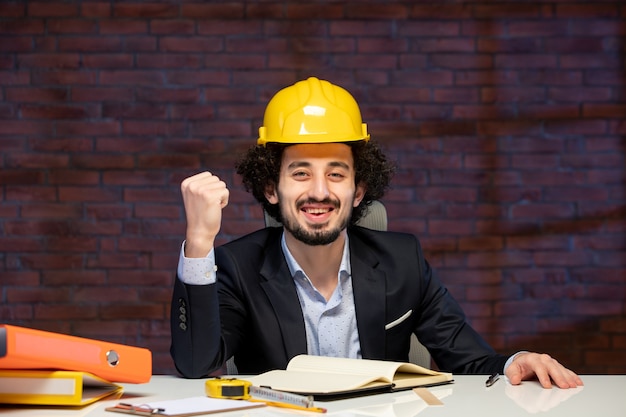 Vorderansicht des männlichen ingenieurs, der hinter dem arbeitsplatz in anzug und gelbem helm sitzt