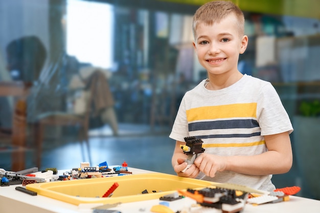 Vorderansicht des reizenden kaukasischen Jungen lächelnd und direkt schauend. Baukasten für Kinder auf dem Tisch, Kinder, die Spielzeug schaffen