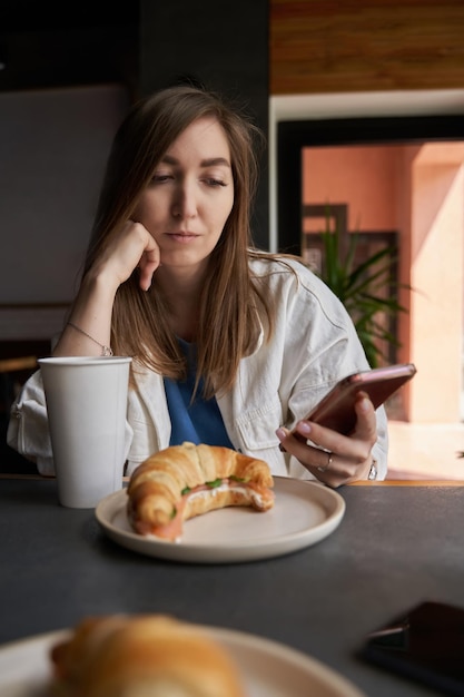 Vorderansicht einer attraktiven Frau, die eine Brille trägt und mit dem Smartphone Kaffee im Café trinkt