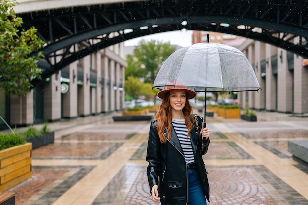 Vorderansicht einer glücklichen jungen Frau mit Modehut, die auf der Straße der europäischen Stadt mit transparentem Regenschirm spazieren geht und lächelnd in die Kamera blickt