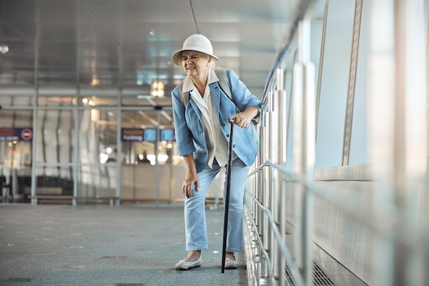 Vorderansicht einer Touristin mit einem Gehstock, die am Flughafen plötzliche Knieschmerzen erleidet