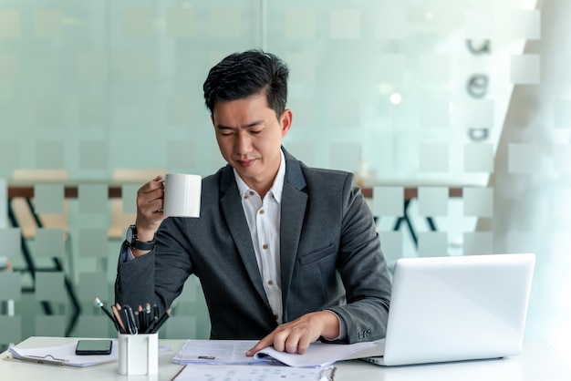 Vorderansicht eines asiatischen Geschäftsmannes, der Kaffee trinkt, während er den im Büro platzierten Dokumentenlaptop überprüft.