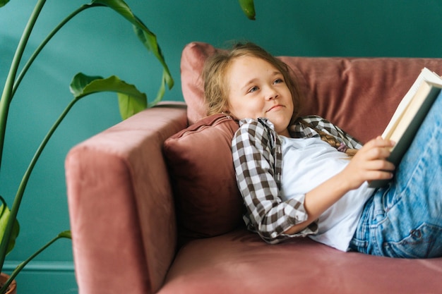 Vorderansicht eines fröhlichen, entzückenden Kindermädchens, das ein Papierbuch liest, das zu Hause auf einer weichen Couch liegt?