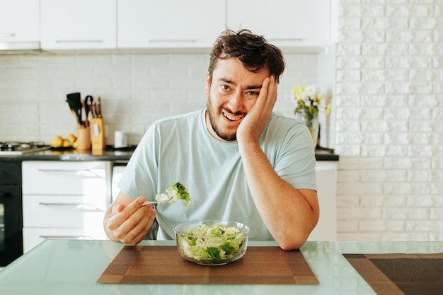 Vorderansicht eines jungen Mannes, der mit unzufriedenem Gesicht an einem Tisch sitzt und einen Salat isst