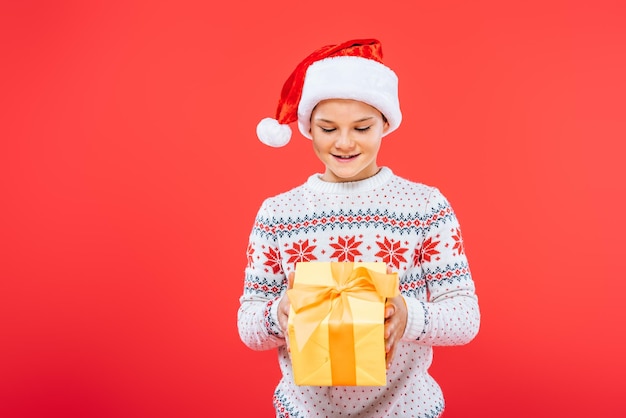 Vorderansicht eines lächelnden Kindes mit Weihnachtsmütze, das ein Geschenk isoliert auf Rot hält