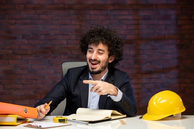 Vorderansicht eines männlichen Ingenieurs, der hinter dem Arbeitsplatz im Anzug des Bauunternehmers sitzt