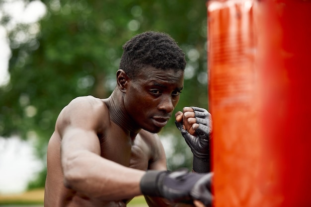 Vorderansicht eines muskulösen schwarzen Boxers, der mit einem tiefen und intensiven Gesicht im Freien auf die Kamera zuschlägt. Box- und Trainingskonzept