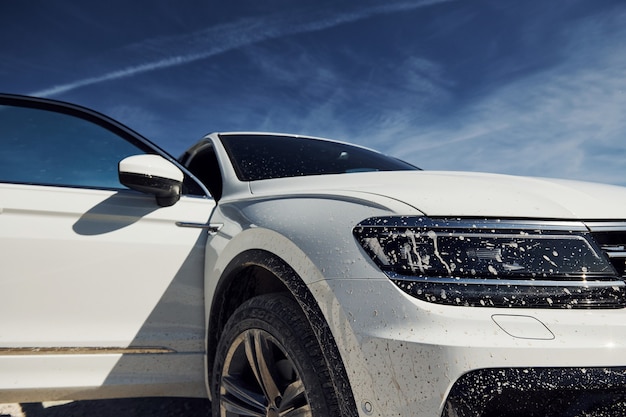 Vorderansicht eines weißen modernen autos, das im freien vor blauem himmel im hintergrund und mit schmutz auf den scheinwerfern geparkt wurde.