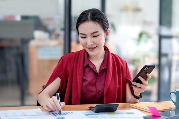Vorderansicht junge asiatische Geschäftsfrau, die ein rotes Kleid trägt und ein Smartphone hält, das mit einem Diagramm im Büro arbeitet.