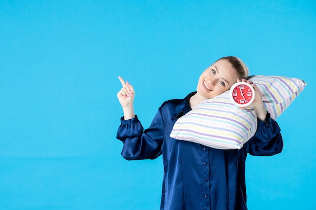 Vorderansicht junge Frau im Pyjama, die Uhren und Kissen auf blauem Hintergrund hält müde Bett Nacht Party Ruhe Schlaf Albtraum späte Frau