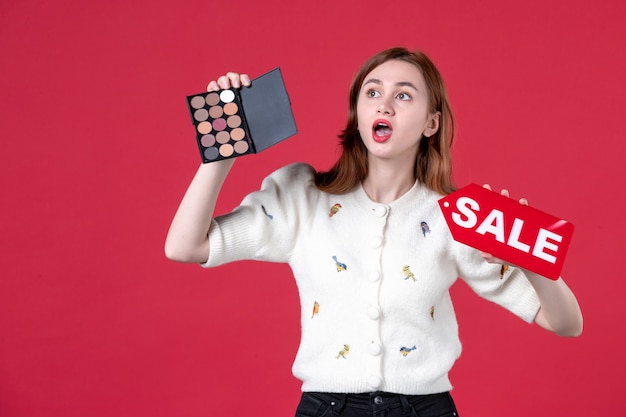Vorderansicht junge Frau mit Puderdose und rotem Verkaufsschild auf rotem Hintergrund sinnliche Einkaufsfrau März Make-up Schönheit Haut Mode Lippen Farben