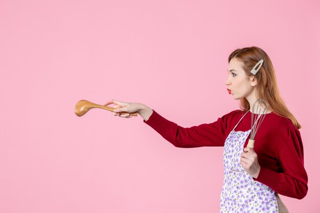 Vorderansicht junge Hausfrau posiert mit Schneebesen und Holzlöffel auf rosa Hintergrund Kuchen Kuchen Beruf Kochen einheitliche Frau Teig horizontale Küche