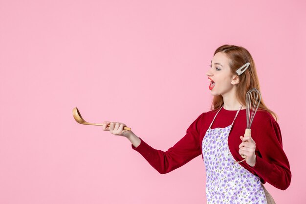 Vorderansicht junge Hausfrau posiert mit Schneebesen und Holzlöffel auf rosa Hintergrund Tortenkuchen kochen einheitliche Frau Teig horizontale Küche