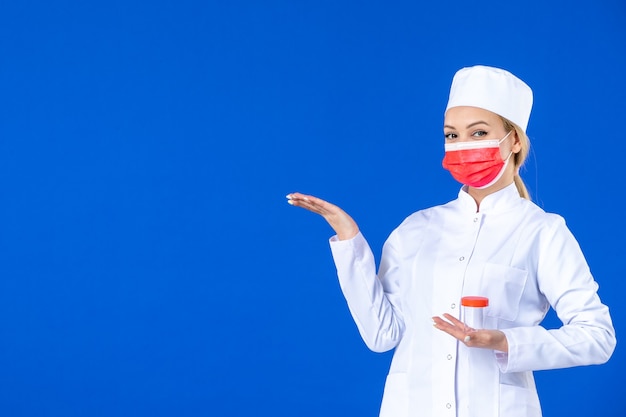 Vorderansicht junge Krankenschwester im medizinischen Anzug mit Maske, die Flasche auf blauem Hintergrund hält Krankenhausvirus Covid-Doktor Impfstoff Pandemie Medizin Medikament