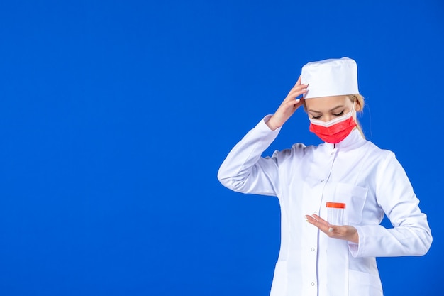 Vorderansicht junge Krankenschwester im medizinischen Anzug mit Maske, die Flasche auf blauem Hintergrund hält