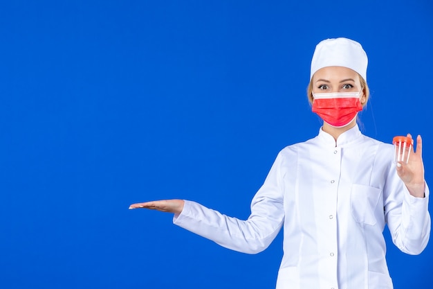Vorderansicht junge Krankenschwester im medizinischen Anzug mit roter Maske, die Flaschen auf blauem Hintergrund hält