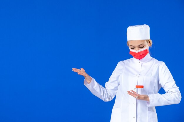 Vorderansicht junge Krankenschwester in medizinischem Anzug mit Maske, die Flasche auf blauem Hintergrund hält Krankenhausvirus Covid-Doktor Impfstoff pandemische Gesundheitsmedizin Medikamente