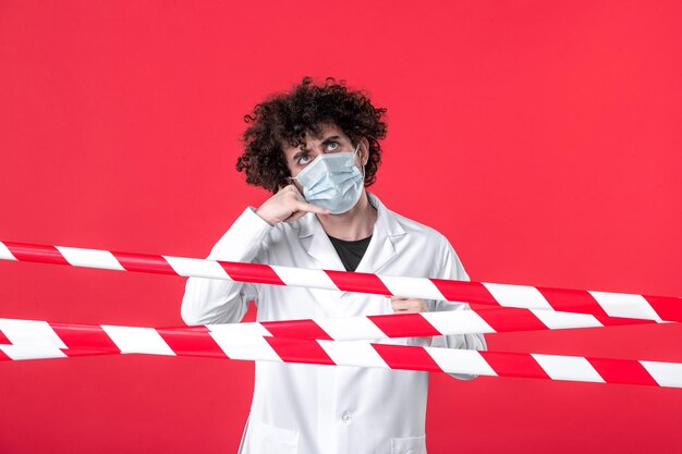Vorderansicht junger männlicher Arzt in medizinischer Uniform und Maske auf rotem Hintergrund Gefahr Isolation Krankenhaus Covid-Gesundheit Quarantäne Warnstreifen