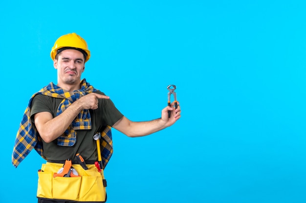 Vorderansicht junger männlicher Baumeister, der eine Zange auf dem blauen Hintergrund hält Konstruktor Architektur Arbeiter Hausbau Farbwerkzeug flach