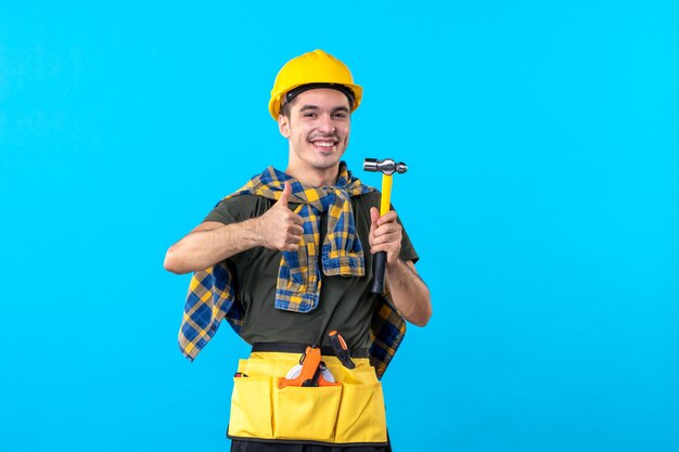 Foto vorderansicht junger männlicher baumeister, der hammer auf blauem hintergrund hält arbeiterhauskonstrukteur flache arbeit gebäudearchitektur