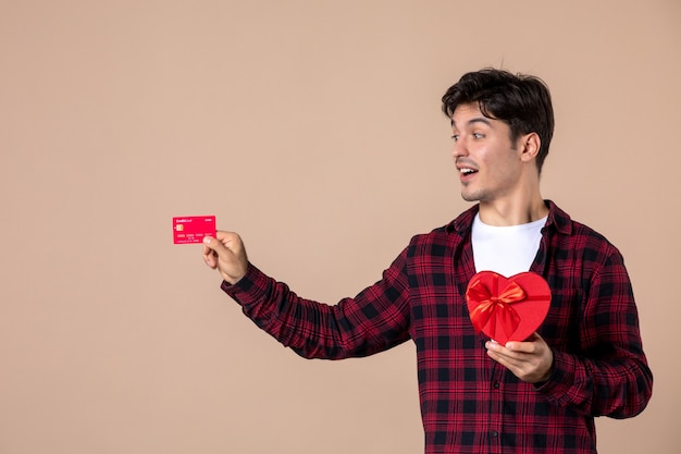 Vorderansicht junger Mann, der herzförmiges Geschenk und Bankkarte an brauner Wand hält