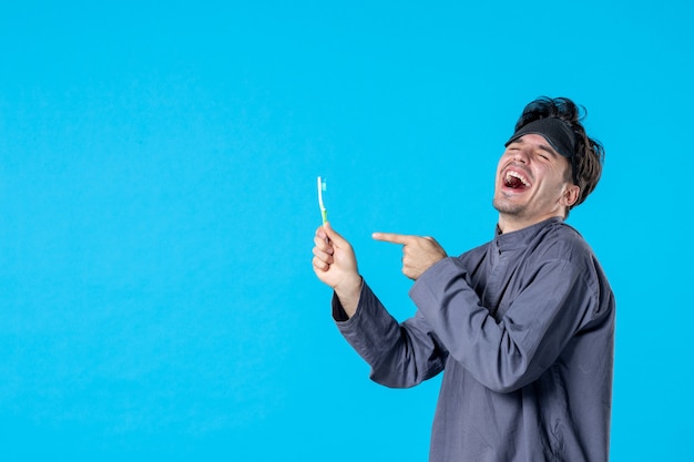Vorderansicht junger Mann im Pyjama, der auf Zahnbürste zeigt und auf blauem Hintergrund lacht Farbe Traum Alptraum Nacht aufwachen sauberes menschliches Bett