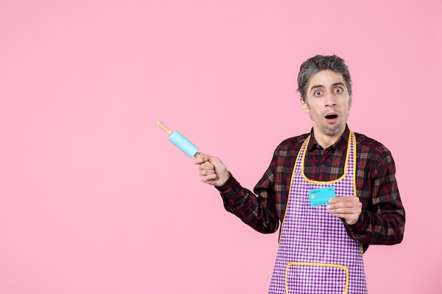Vorderansicht junger Mann im Umhang mit kleinem Nudelholz und Bankkarte auf rosa Hintergrund Ehemann Arbeiter Uniform Geld horizontaler Job Küche Kochen