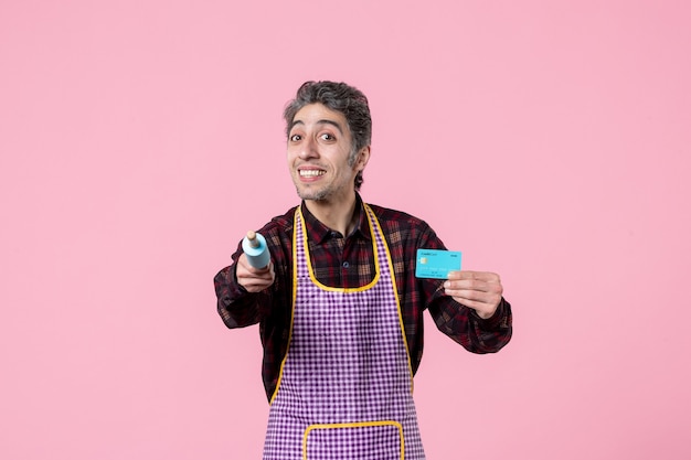 Foto vorderansicht junger mann im umhang mit kleinem nudelholz und bankkarte auf rosa hintergrund einheitliche geldküche beruf kochen ehemann horizontale arbeit