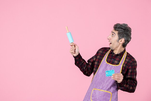 Vorderansicht junger Mann im Umhang mit kleinem Nudelholz und Bankkarte auf rosa Hintergrund Job Ehemann Arbeiter Uniform Beruf Küche Kochen Geld