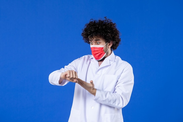 Vorderansicht nervöser männlicher arzt in medizinischem anzug und maske, die zeit auf blauem hintergrund überprüft