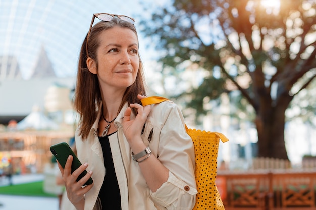 Vorderansicht Porträt moderne Mode glückliche Frau Hipster zu Fuß und mit einem Smartphone auf einer Stadtstraße mit Sonnenbrille