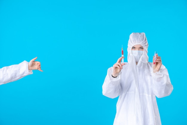 Vorderansicht Ärztin in Schutzanzug und Maske mit Injektion auf blauem Hintergrund medizinisches Krankenhaus covid pandemische Gesundheitswissenschaft