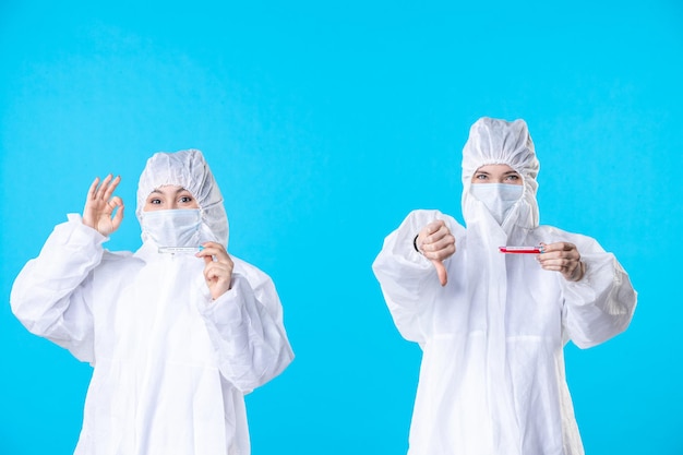 Vorderansicht Ärztinnen in Schutzanzügen und Maske mit Flaschen auf blauem Hintergrund Viruspandemie Wissenschaft Covid-Krankenhaus